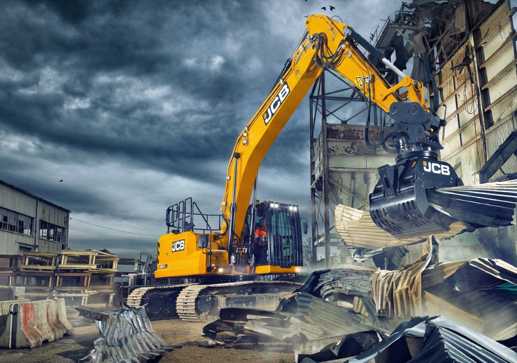 JCB-merkkinen kaivinkone, joka on varustettu purkukouralla, on juuri nostamassa tai repimässä irti suurta kasaa aaltopeltiä ja muuta rakennusjätettä.