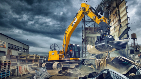 JCB-merkkinen kaivinkone, joka on varustettu purkukouralla, on juuri nostamassa tai repimässä irti suurta kasaa aaltopeltiä ja muuta rakennusjätettä.